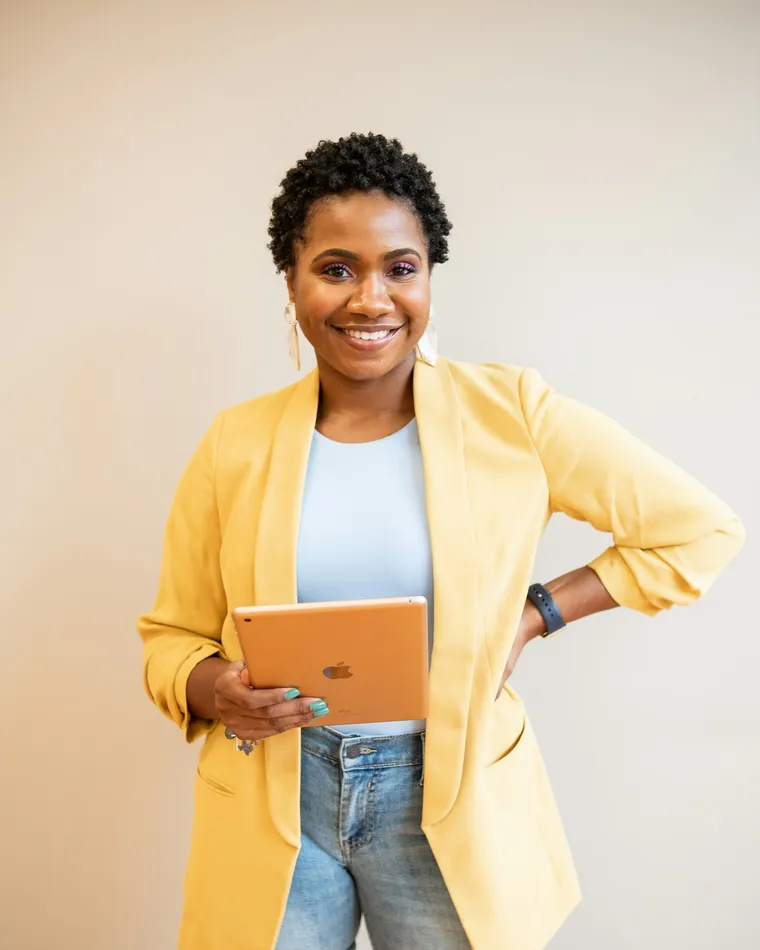 A smiling woman in a yellow blazer holding a tablet